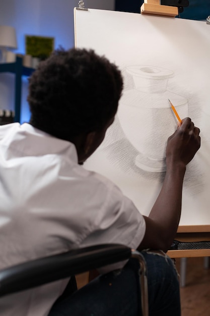 Afroamerikanischer Künstler im Rollstuhl, der Schatten mit grafischem Bleistift macht, der während der Malstunde im Kunstatelier an der Vasenillustration arbeitet. Kreativer Student mit Behinderung, der eine künstlerische Idee skizziert