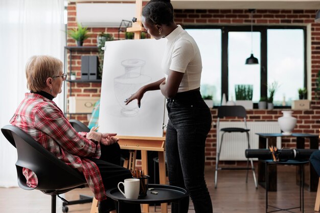 Afroamerikanischer Kunstlehrer erklärt älteren Schülern während des Kreativitätskurses die Zeichentechnik. Künstlerin skizziert Vasenmodell auf weißer Leinwand mit grafischem Bleistift. Neujahrsvorsätze