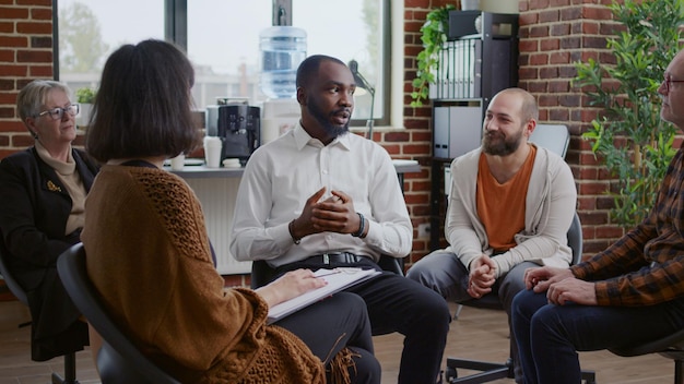 Afroamerikanischer Mann, der die Suchtgeschichte mit einer Gruppe von Menschen bei einem Therapietreffen teilt. Erwachsene im Gespräch mit Psychologen und Patienten im Kreis bei der Rehabilitationssitzung.