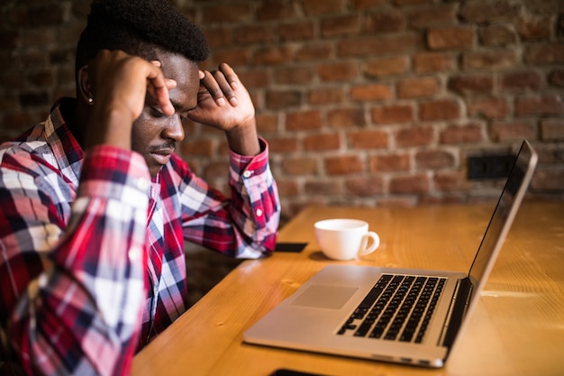 Afroamerikanischer Mann, der Kopfschmerzen während der Fernarbeit im Coffeeshop fühlt, müde vom Versagen von Plänen, männlicher Unternehmer überarbeitet, um Probleme mit Startup-Projekt mit hartem Zeitplan erschöpft zu lösen