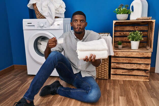 Afroamerikanischer Mann hält saubere Wäsche und Waschpulver in der Hand und macht das Fischgesicht mit Mund und schielenden Augen verrückt und komisch