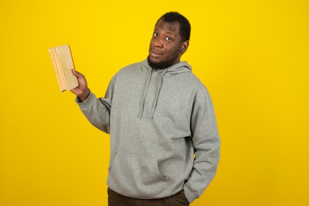 Afroamerikanischer Mann mit grauem Sweatshirt, mit einem Buch in der einen Hand und der anderen in der Tasche, steht über der gelben Wand.