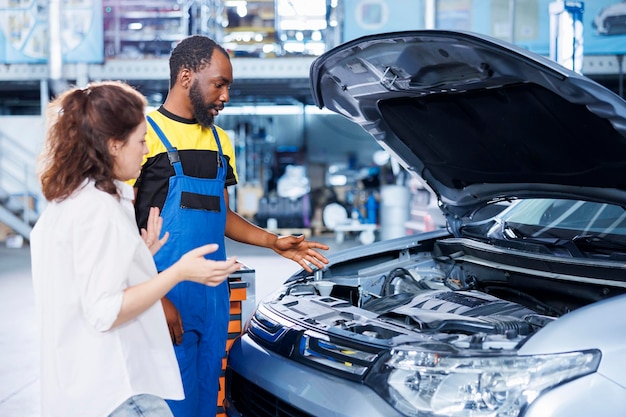 Afroamerikanischer Mechaniker hilft einem Kunden bei der Wartung von Autos in einer Autowerkstatt Angestellter in einer Garage, der Autoteile mit einer Frau repariert, die während der Inspektion ihren Fahrzeugmotor repariert