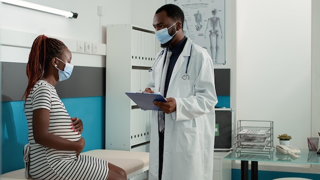 Afroamerikanischer Mediziner und schwangere Patientin treffen sich nach Terminvereinbarung während der Coronavirus-Pandemie. Arzt spricht mit Frau mit Schwangerschaftsbauch und gibt ärztlichen Rat beim Kontrollbesuch.