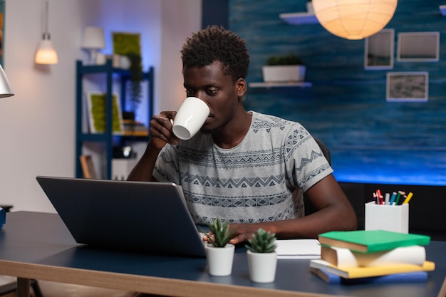 Afroamerikanischer Mitarbeiter, der eine Tasse Kaffee hält und Marketingideen auf dem Computer eingibt, der bei einem Online-Projekt im Wohnzimmer remote arbeitet. Freiberufler, der am Schreibtisch sitzt und Finanzdiagramm analysiert