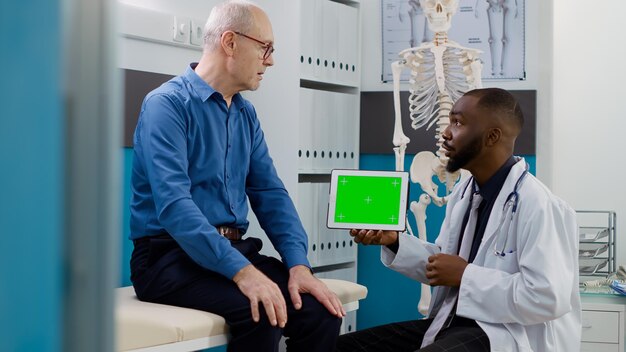 Afroamerikanischer Sanitäter hält Tablet mit horizontalem Greenscreen bei der Untersuchung mit älterem Patienten. Arzt und Mann analysieren isolierten Chromakey-Kopienraum mit leerer Mockup-Vorlage auf dem Display.