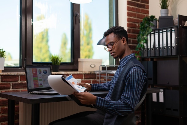 Afroamerikanischer Startup-Besitzer mit Brille, die Klemmbrett und Papier mit Geschäftsdiagrammen hält, die Daten mit Verkaufsergebnissen auf dem Laptop vergleichen. Unternehmer, der Marketingindikatoren auf Laptop analysiert.