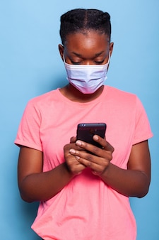 Afroamerikanischer student mit schützender medizinischer gesichtsmaske gegen coronavirus, der ein smartphone hält und mit freunden im studio mit blauem hintergrund nachrichten schreibt. frau gibt text ein