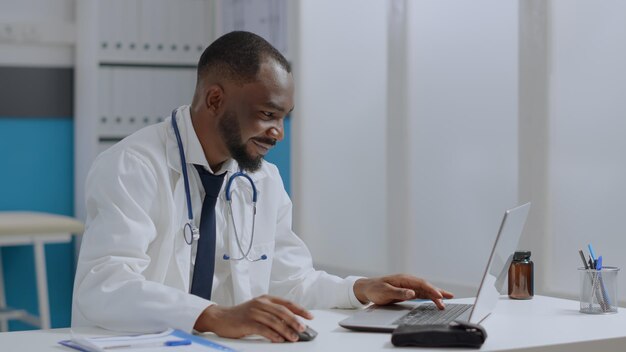 Afroamerikanischer Therapeut, Arzt, der am Schreibtisch sitzt und Krankheitsexpertise auf einem Laptop eingibt, der bei der Gesundheitsbehandlung im Krankenhausbüro arbeitet. Arztmann analysiert Krankheitsbericht. Medizin-Konzept