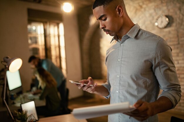 Afroamerikanischer Unternehmer liest Nachricht auf Smartphone, während er nachts im Büro an Geschäftsberichten arbeitet