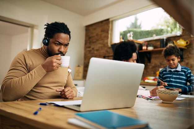 Afroamerikanischer Vater trinkt Tee, während er zu Hause am Laptop arbeitet