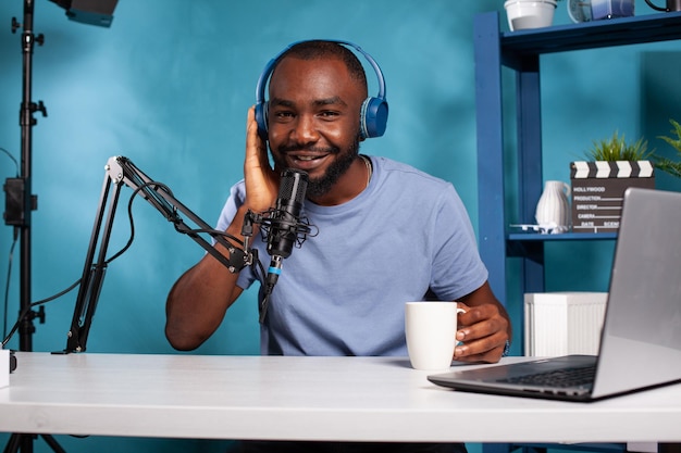 Afroamerikanischer Vlogger, der drahtlose Kopfhörer ans Ohr drückt, während er mit der Hand dem Fan zuhört, der spricht, während er eine weiße Tasse hält. Influencer sitzt am Schreibtisch mit Laptop und interagiert mit dem Publikum.