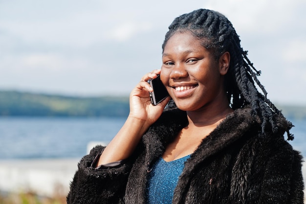 Afroamerikanisches, dunkelhäutiges Plus-Size-Model, das in einem blau glänzenden Kleid und einem schwarzen Pelzmantel vor dem Meer posiert, spricht am Telefon