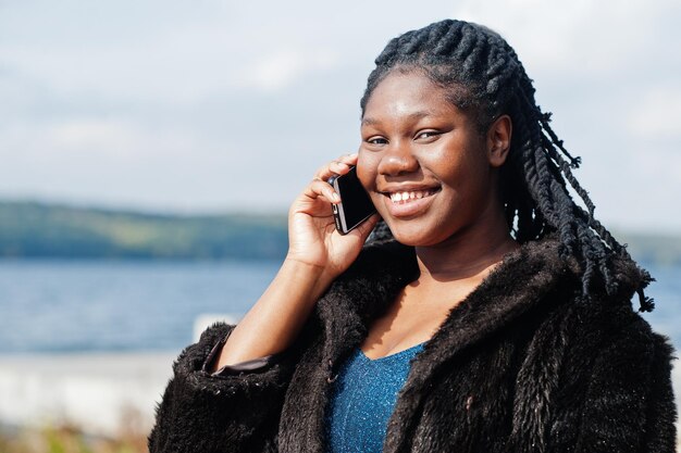 Afroamerikanisches, dunkelhäutiges Plus-Size-Model, das in einem blau glänzenden Kleid und einem schwarzen Pelzmantel vor dem Meer posiert, spricht am Telefon