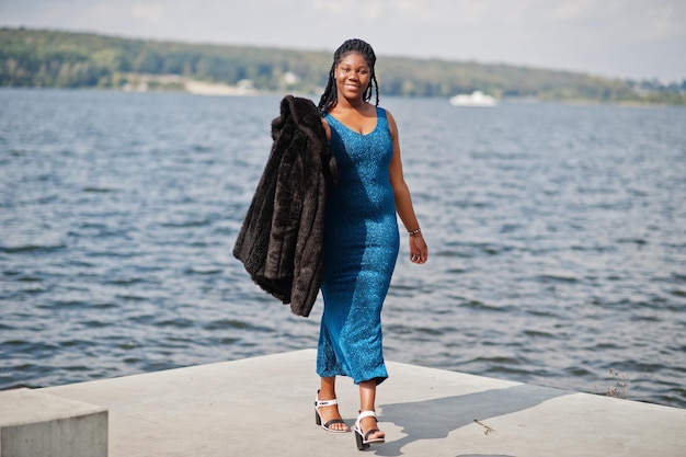 Afroamerikanisches, dunkelhäutiges Plus-Size-Model posierte in einem blau glänzenden Kleid und schwarzem Pelzmantel an der Meeresseite