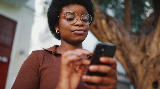 Afroamerikanisches lockiges Mädchen mit Brille unter Verwendung eines Smartphones f