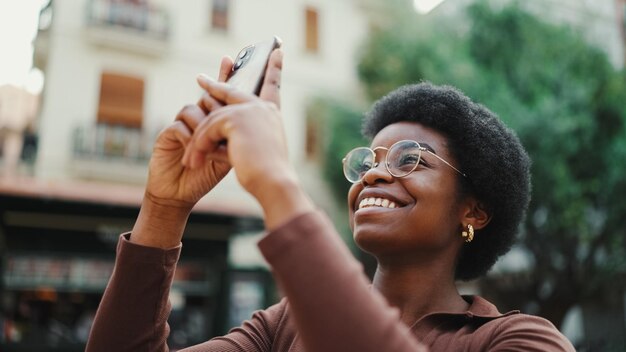Afroamerikanisches Mädchen, das Foto von schöner Architektur am Telefon im Freien macht Fröhliche Frau mit Brille, die auf der Straße lächelt