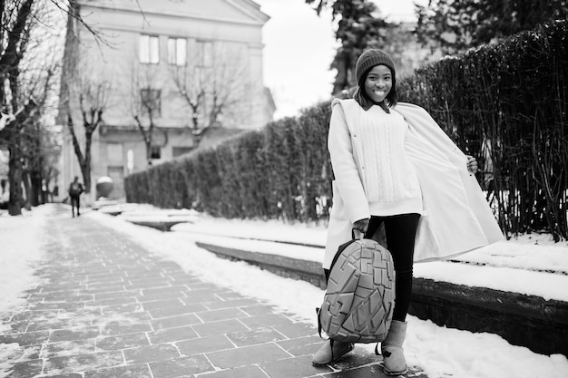 Afroamerikanisches Mädchen mit rotem Hut und rosafarbenem Mantel mit Rucksack auf der Straße der Stadt am Wintertag