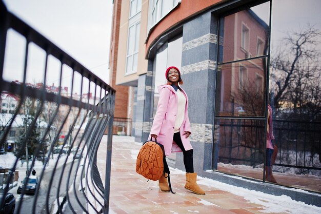 Afroamerikanisches Mädchen mit rotem Hut und rosafarbenem Mantel mit Rucksack auf der Straße der Stadt gegen Gebäude am Wintertag