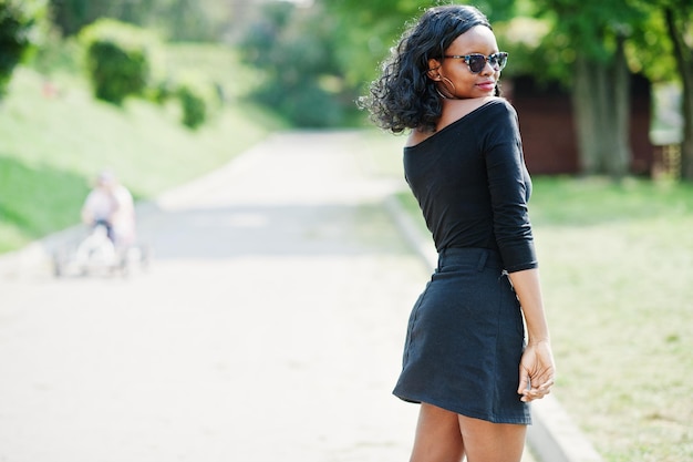Afroamerikanisches Mädchen mit Sonnenbrille, schwarzer Kleidung und Hemd posierte im Freien Modische schwarze Frau