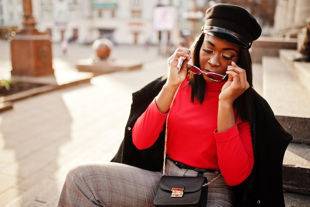 Afroamerikanisches Modemädchen in Mantel- und Zeitungsjungenkappen-Sonnenbrille posierte auf der Straße