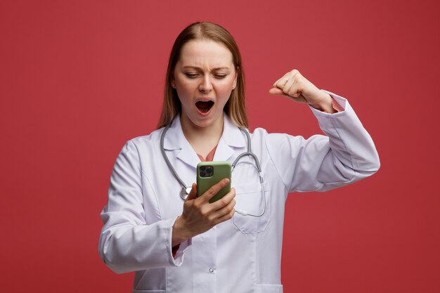 Aggressive junge blonde Ärztin, die medizinische Robe und Stethoskop um den Hals hält, hält und betrachtet Handy, das starke Geste tut