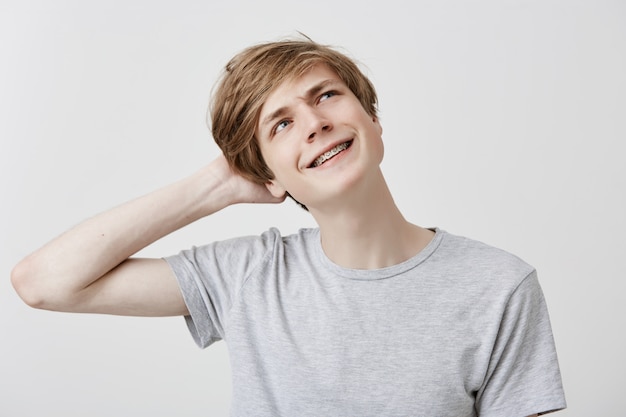 Ahnungslos verwirrter junger kaukasischer Mann im grauen T-Shirt mit hellem Haar und blauen Augen, die mit verwirrtem und verwirrtem Ausdruck nach oben schauen, Kopf kratzen, den Geburtstag der Freundin vergessen