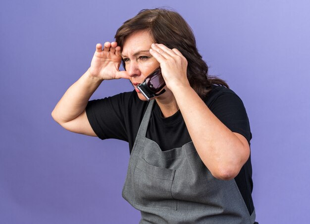 Ahnungslose erwachsene kaukasische Friseurin in Uniform, die die Handfläche an der Stirn hält und die Haarschneidemaschine mit Blick auf die Seite isoliert auf violettem Hintergrund mit Kopienraum hält