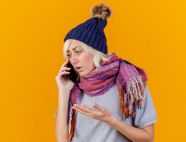 Ahnungslose junge blonde kranke slawische Frau, die Wintermütze und Schal trägt, spricht am Telefon, das Seite betrachtet