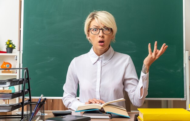 Ahnungslose junge blonde Lehrerin mit Brille, die am Schreibtisch mit Schulwerkzeugen im Klassenzimmer sitzt und die Hand auf offenem Buch hält und auf die Kamera blickt, die leere Hand zeigt