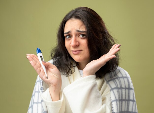 Ahnungslose junge kranke Frau, die Robe wickelt, die im karierten Haltethermometer wickelt, das Front betrachtet, die leere Hand lokalisiert auf olivgrüner Wand zeigt