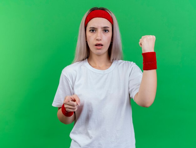 Ahnungslose junge sportliche Frau mit Zahnspangen, die Stirnband und Armbänder tragen, hält Faust und Punkte vorne isoliert auf grüner Wand