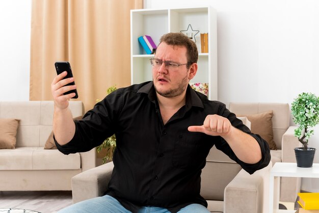 Ahnungsloser erwachsener slawischer Mann mit optischer Brille sitzt auf einem Sessel und schaut auf das Telefon