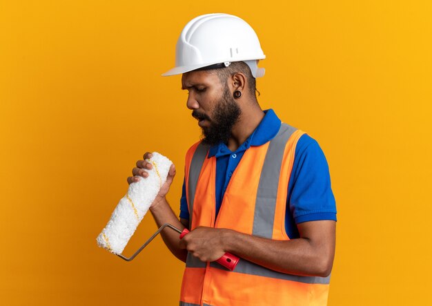 Ahnungsloser junger afroamerikanischer Baumeister in Uniform mit Schutzhelm, der Farbroller einzeln auf orangefarbenem Hintergrund mit Kopierraum hält und betrachtet