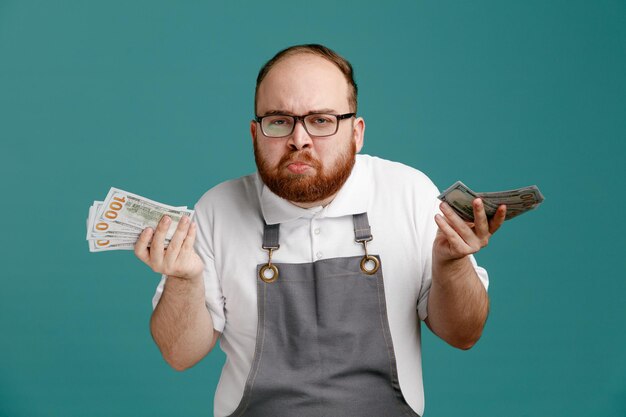 Ahnungsloser junger Friseur in Uniform und Brille mit Geld in beiden Händen, der isoliert auf blauem Hintergrund in die Kamera blickt