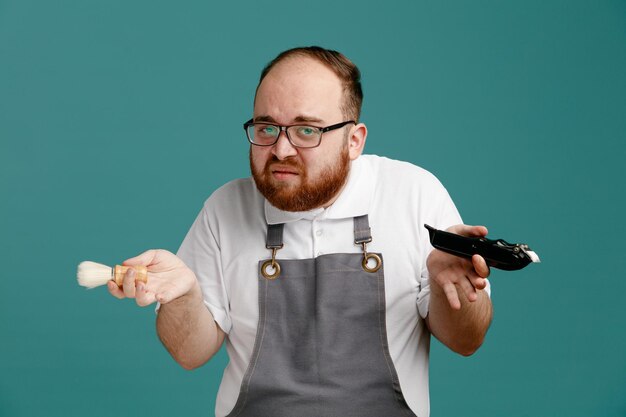 Ahnungsloser junger Friseur in Uniform und Brille mit Rasierpinsel und Haarschneider, der isoliert auf blauem Hintergrund in die Kamera blickt