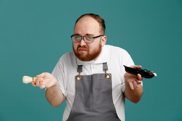 Ahnungsloser junger Friseur in Uniform und Brille mit Rasierpinsel und Haarschneider, der isoliert auf blauem Hintergrund in die Kamera blickt