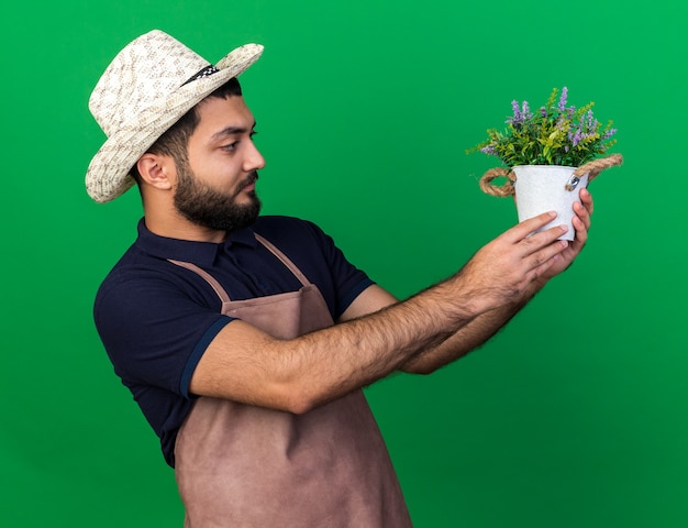 ahnungsloser junger kaukasischer männlicher Gärtner mit Gartenhut, der Blumentopf isoliert auf grüner Wand mit Kopienraum hält und betrachtet