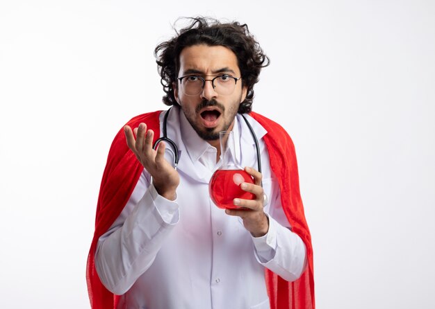 Ahnungsloser junger kaukasischer Mann in der optischen Brille, die Arztuniform mit rotem Umhang und mit Stethoskop um Hals trägt, steht mit erhobener Hand und hält rote chemische Flüssigkeit im Glaskolben
