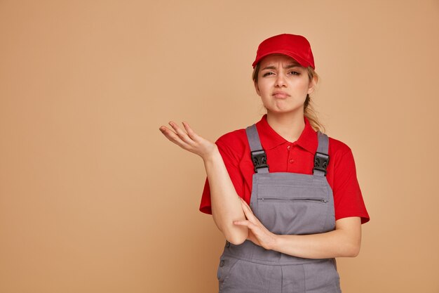 Ahnungsloser junger weiblicher Bauarbeiter, der Kappe und Uniform trägt, die leere Hand zeigen