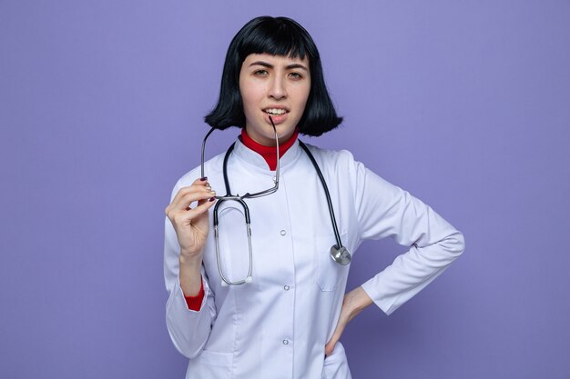 Ahnungsloses junges hübsches kaukasisches Mädchen in Arztuniform mit Stethoskop, das eine optische Brille hält und nach vorne schaut