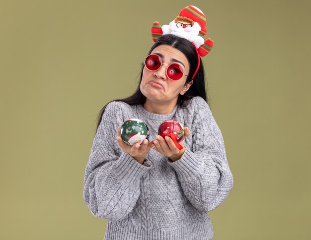 Ahnungsloses junges kaukasisches Mädchen, das Weihnachtsmann-Stirnband mit Brille hält, die Weihnachtskugeln hält, die Kamera lokalisiert auf olivgrünem Hintergrund betrachten