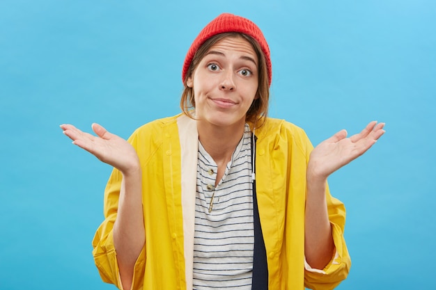 Kostenloses Foto ahnungsloses junges mädchen mit rotem, stilvollem hut und gelbem regenmantel, das mit großen augen auf ihrem gesicht starrt und vor unwissenheit oder gleichgültigkeit mit den schultern zuckt und sagt: dinge passieren