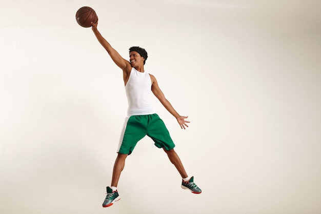 Aktionsfoto eines glücklichen jungen schwarzen Athleten, der weißes Hemd und grüne Shorts trägt, die hoch springen, um einen Weinlesebasketball auf Weiß zu ergreifen