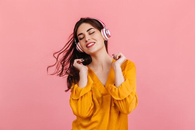 Aktive Frau mit schneeweißem Lächeln tanzt auf rosa Wand. Modell in der orangefarbenen Bluse, die Musik hört.