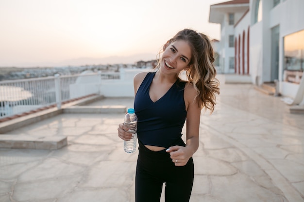 Aktive schlanke junge Frau im Sportbekleidungslauf, lächelnd am Meer bei Sonnenaufgang. Modisches Modell, Training, Training mit Wasser, Lachen, Glück