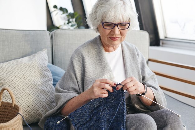 Aktive talentierte kaukasische Frau am Ruhestandstag drinnen verbringen, warme Kleidung für Enkel stricken, auf Sofa in gemütlichem Interieur sitzen, lächelnd. Handarbeit, Handwerk und Hobbykonzept
