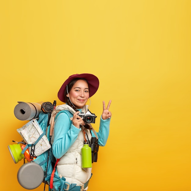 Kostenloses Foto aktive weibliche reisende macht friedensgeste, hält retro-kamera zum fotografieren, trägt großen rucksack mit zielkarte, karemat und anderen touristischen dingen