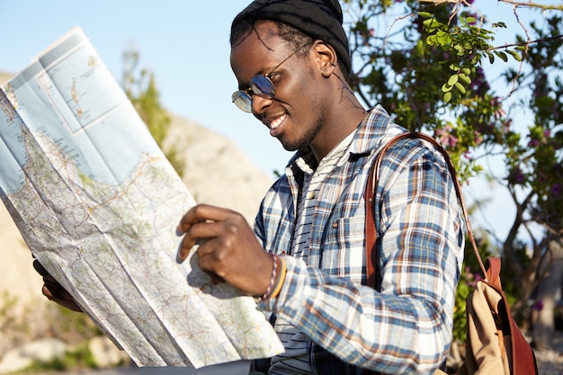Aktiver Lebensstil, Reisen und Tourismus. Fröhlicher modischer junger dunkelhäutiger Reisender mit Rucksack, der Karte hält, die über Straßenfahrt im Berggebiet in der Naturumgebung aufgeregt fühlt