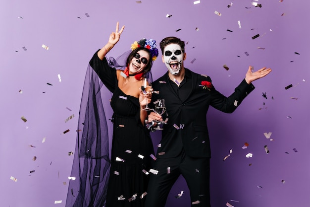 Aktiver Mann und Frau in Kostümen für Halloween tanzen auf lila Hintergrund unter Konfetti.